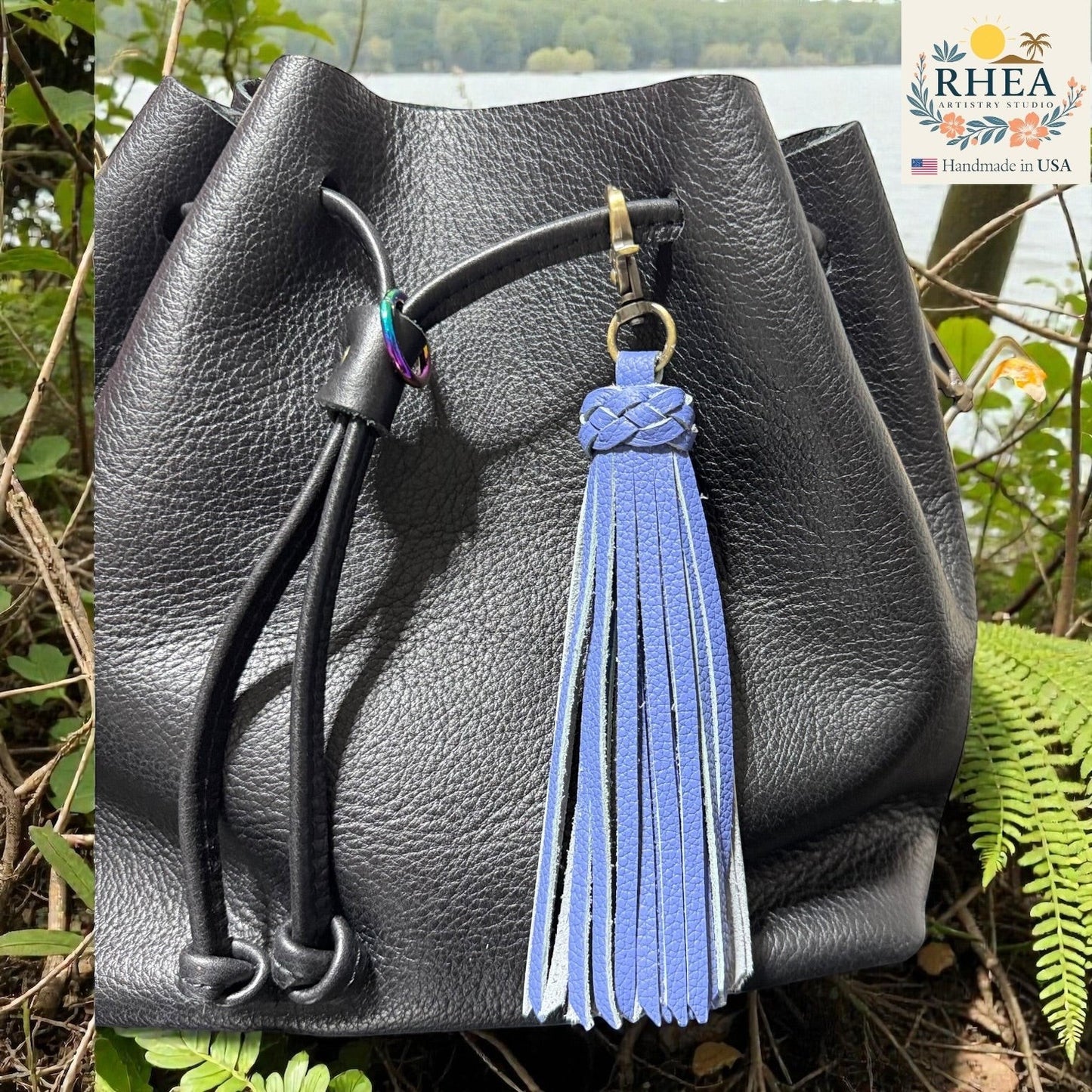 Rhea Artistry Studio blue leather tassel bag charm with braided detail and brass clasp, attached to a black leather bag.