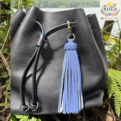 Rhea Artistry Studio blue leather tassel bag charm with braided detail and brass clasp, attached to a black leather bag.