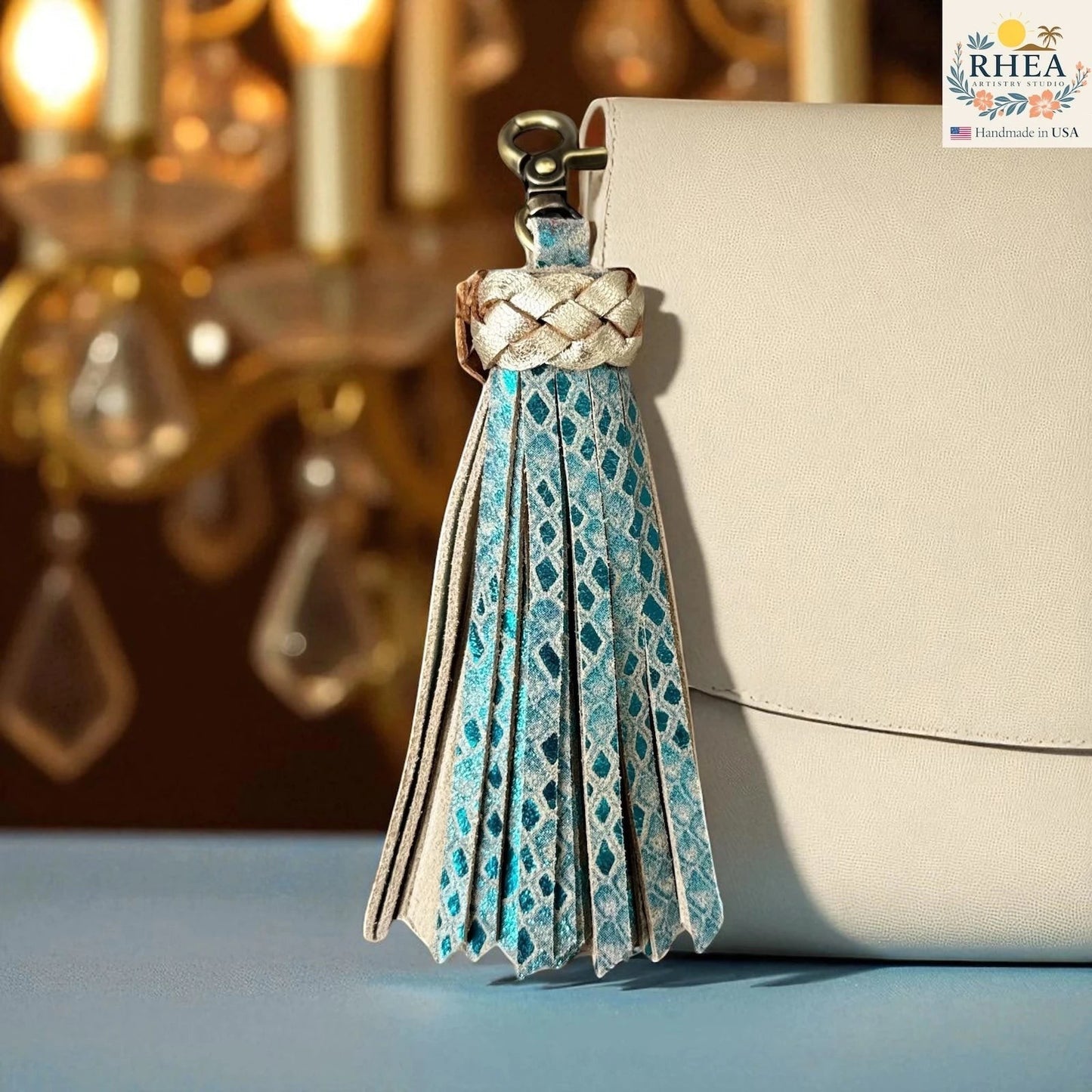 Rhea Artistry Studio handmade leather tassel keychain in blue with diamond pattern and gold braided top, attached to a cream bag.