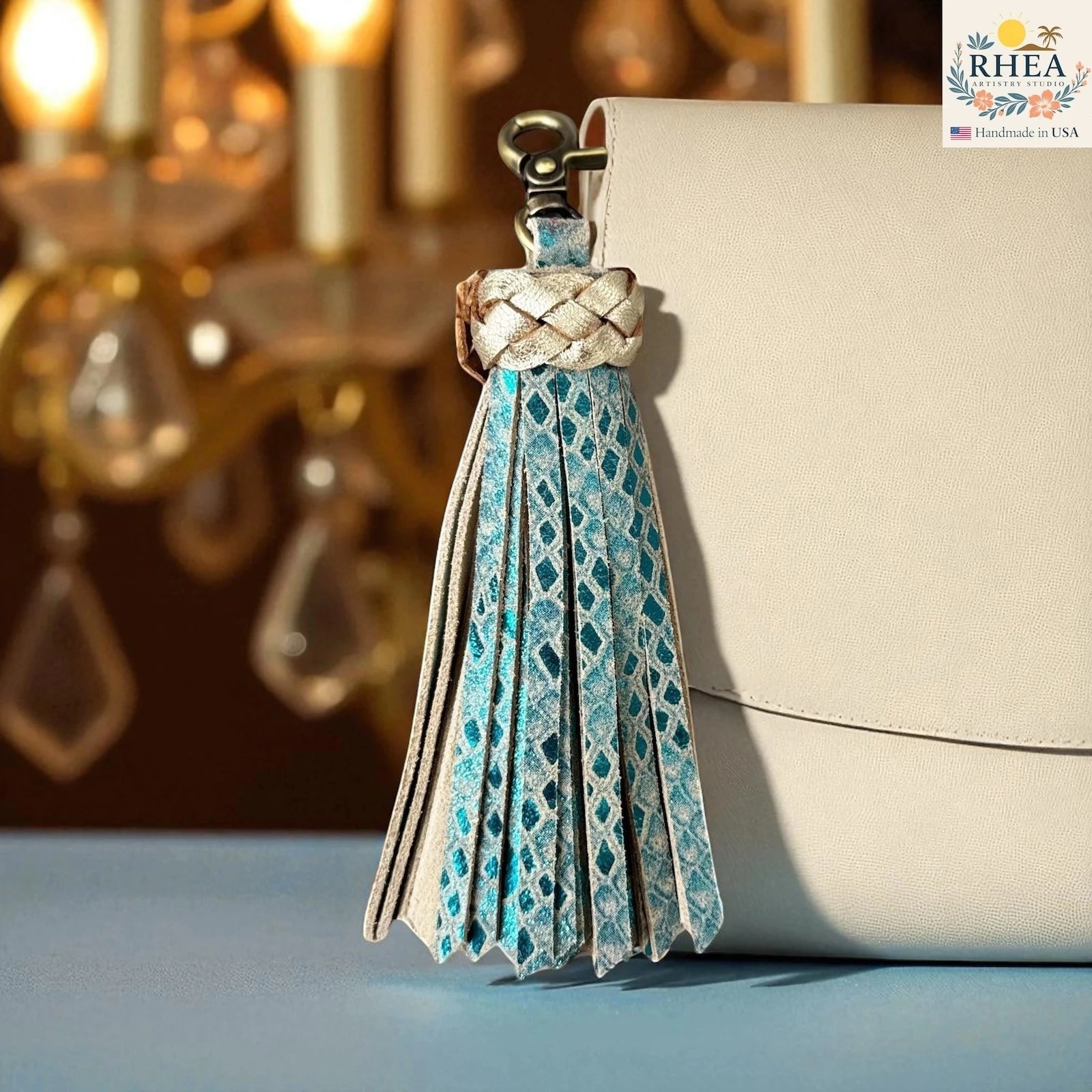Rhea Artistry Studio handmade leather tassel keychain in blue with diamond pattern and gold braided top, attached to a cream bag.