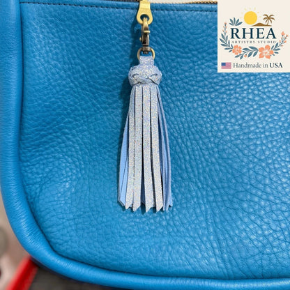 Rhea Artistry Studio handmade light blue leather tassel keychain with glitter accents and antique brass lobster clasp attached to a teal leather bag.