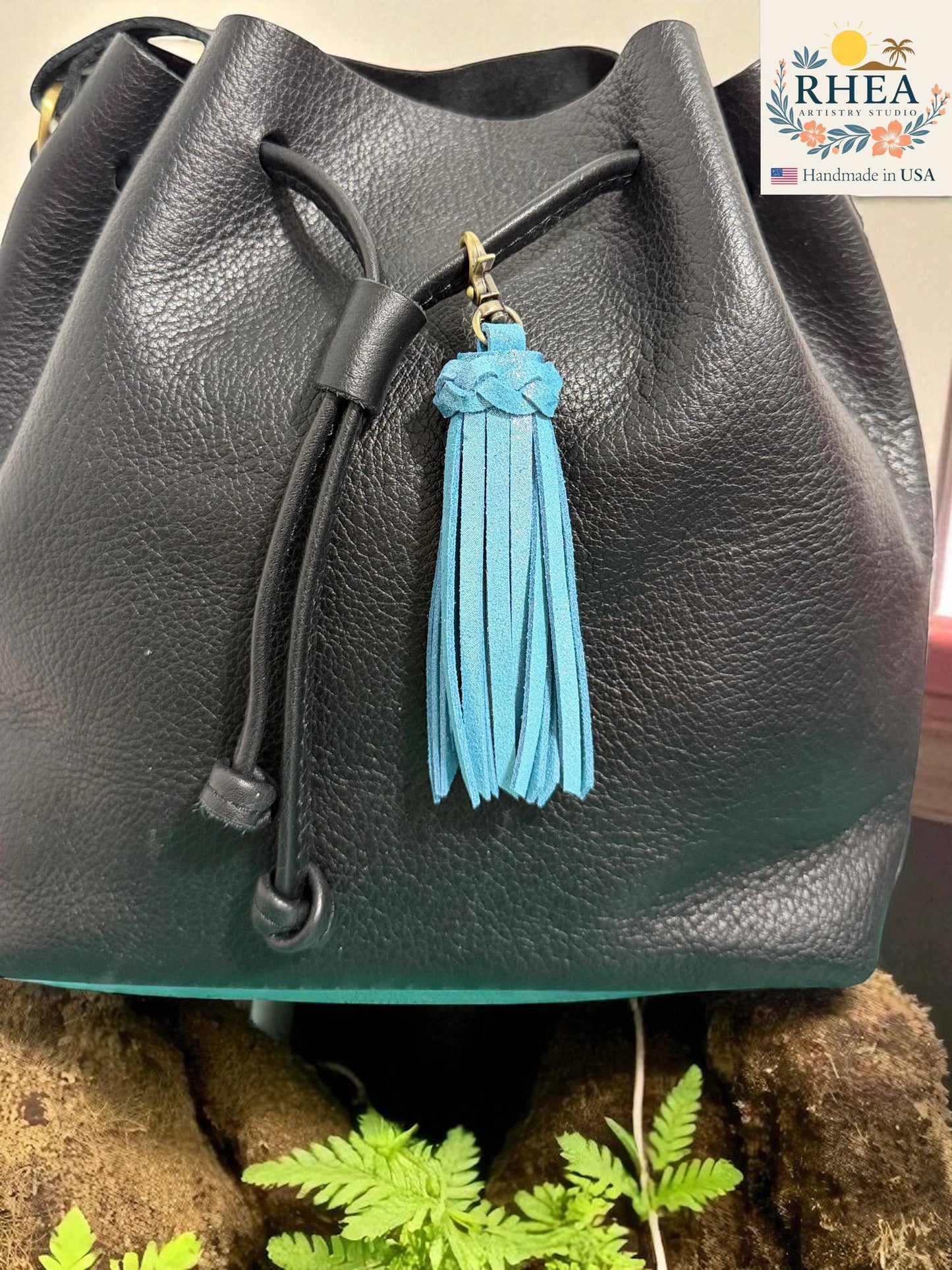 Rhea Artistry Studio handmade bright blue leather tassel keychain with antique brass lobster clasp attached to a black leather bucket bag.