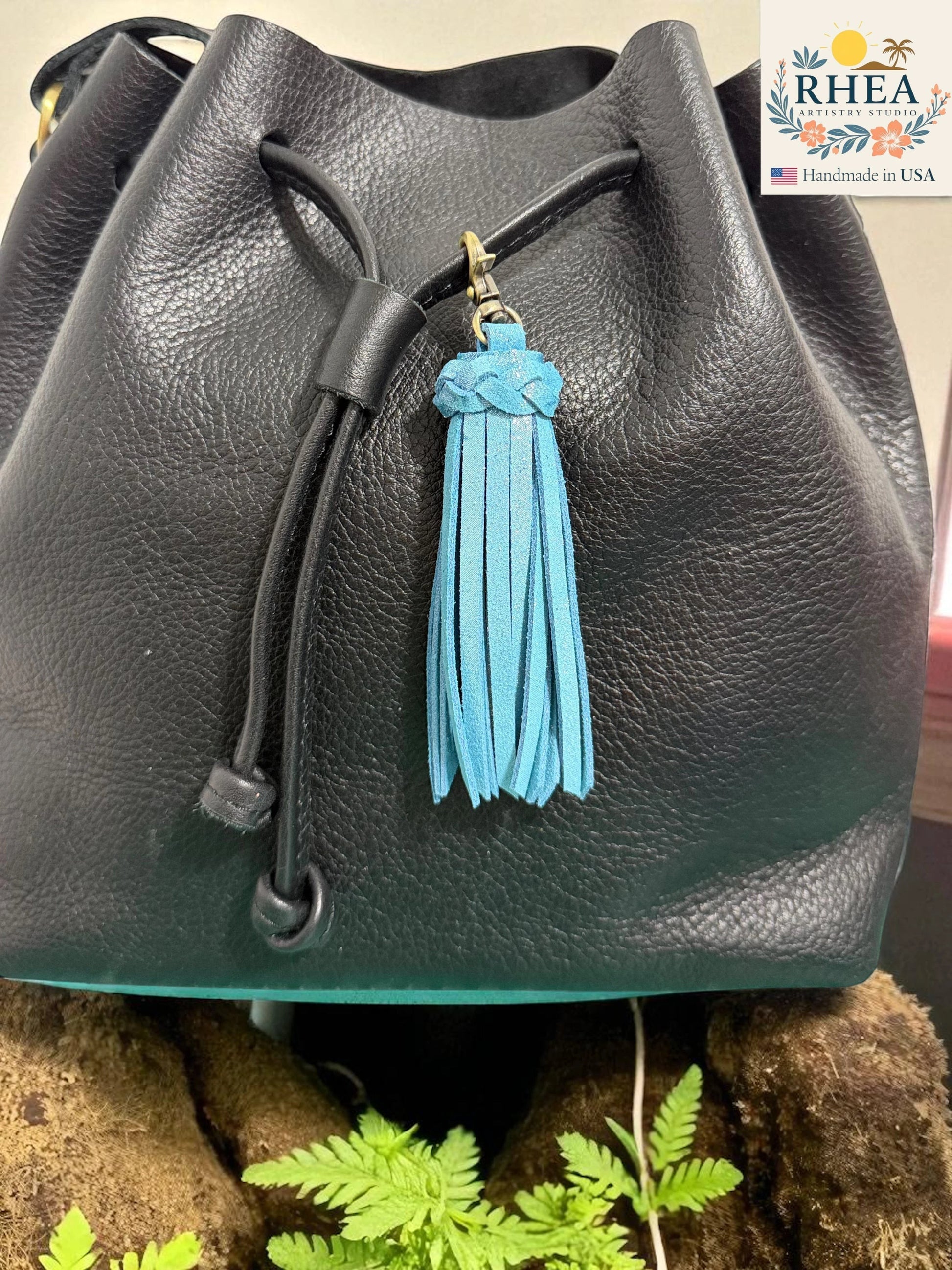 Rhea Artistry Studio handmade bright blue leather tassel keychain with antique brass lobster clasp attached to a black leather bucket bag.