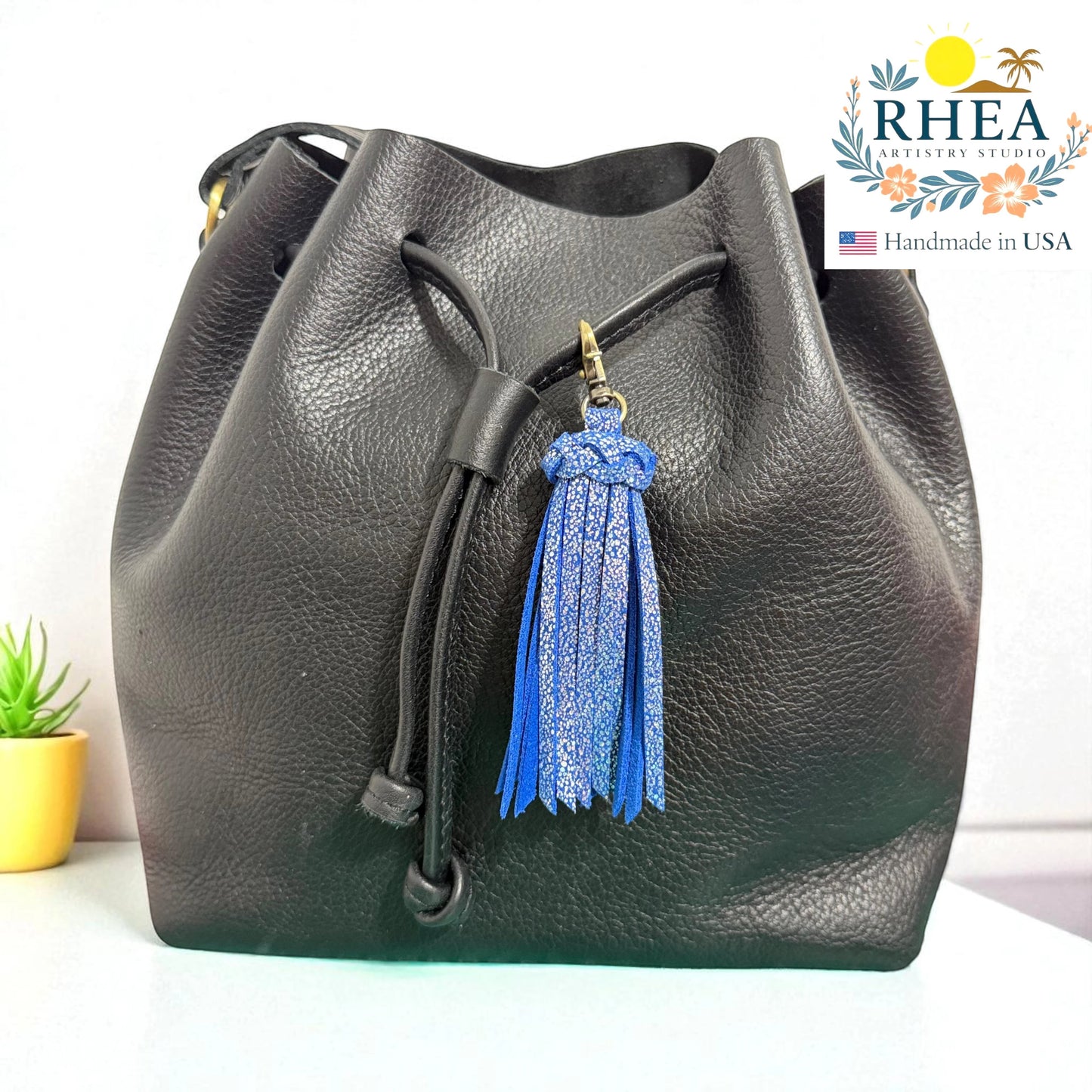 Rhea Artistry Studio blue sparkle leather tassel keychain with antique brass lobster clasp attached to a black leather drawstring bag.