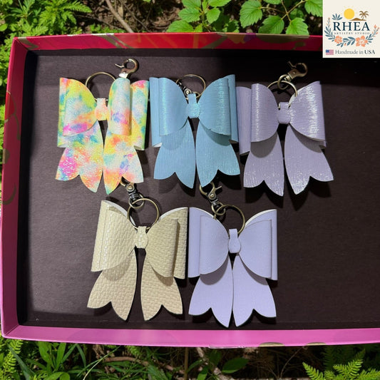 Rhea Artistry Studio handmade double-layer French bow bag charms in pastel blue, lavender, tie-dye glitter, and cream.