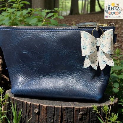 Rhea Artistry Studio handmade double-layer French bow charm with blue bunny print and brass hardware attached to a navy blue leather bag.