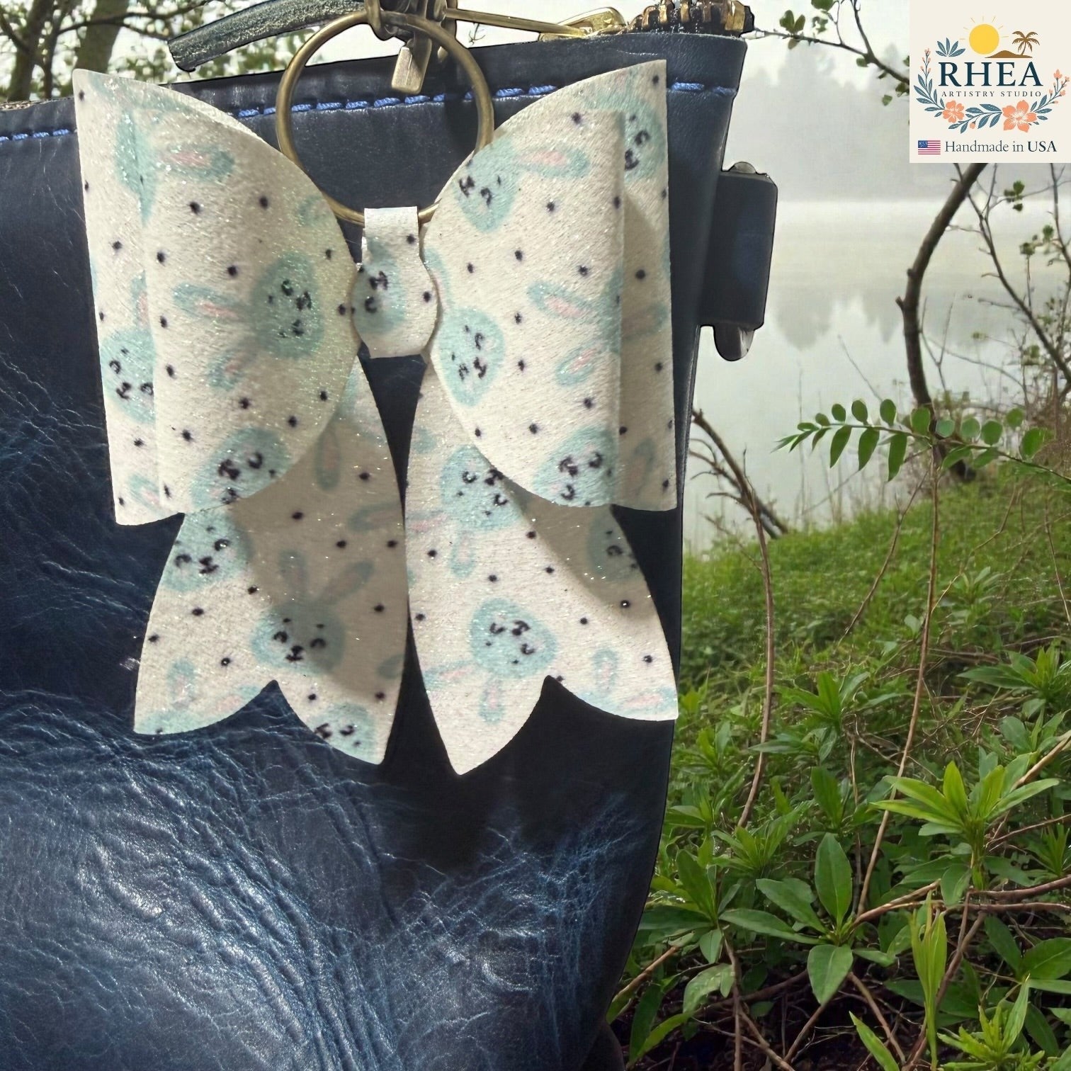 Rhea Artistry Studio handmade white and blue bunny print double-layer French bow bag charm with holographic glitter and brass hardware.
