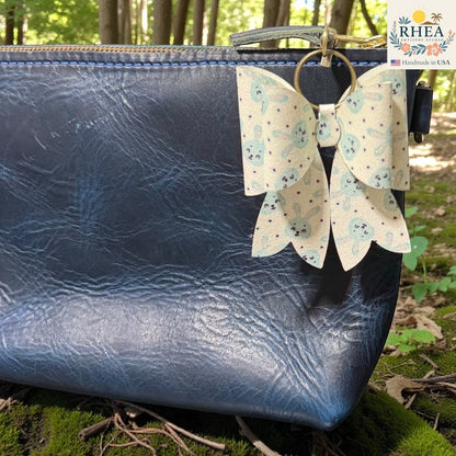 Rhea Artistry Studio handmade double-layer French bow charm with blue bunny print and holographic glitter, attached to a navy blue leather bag.