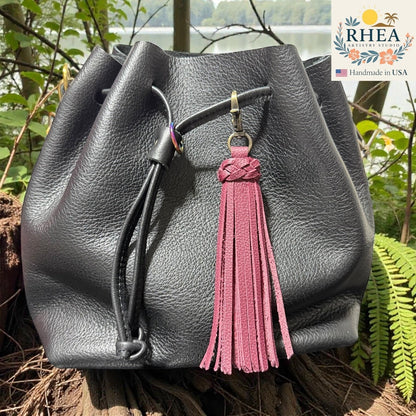 Rhea Artistry Studio burgundy leather tassel bag charm with braided detail and brass clasp on a black leather bag.