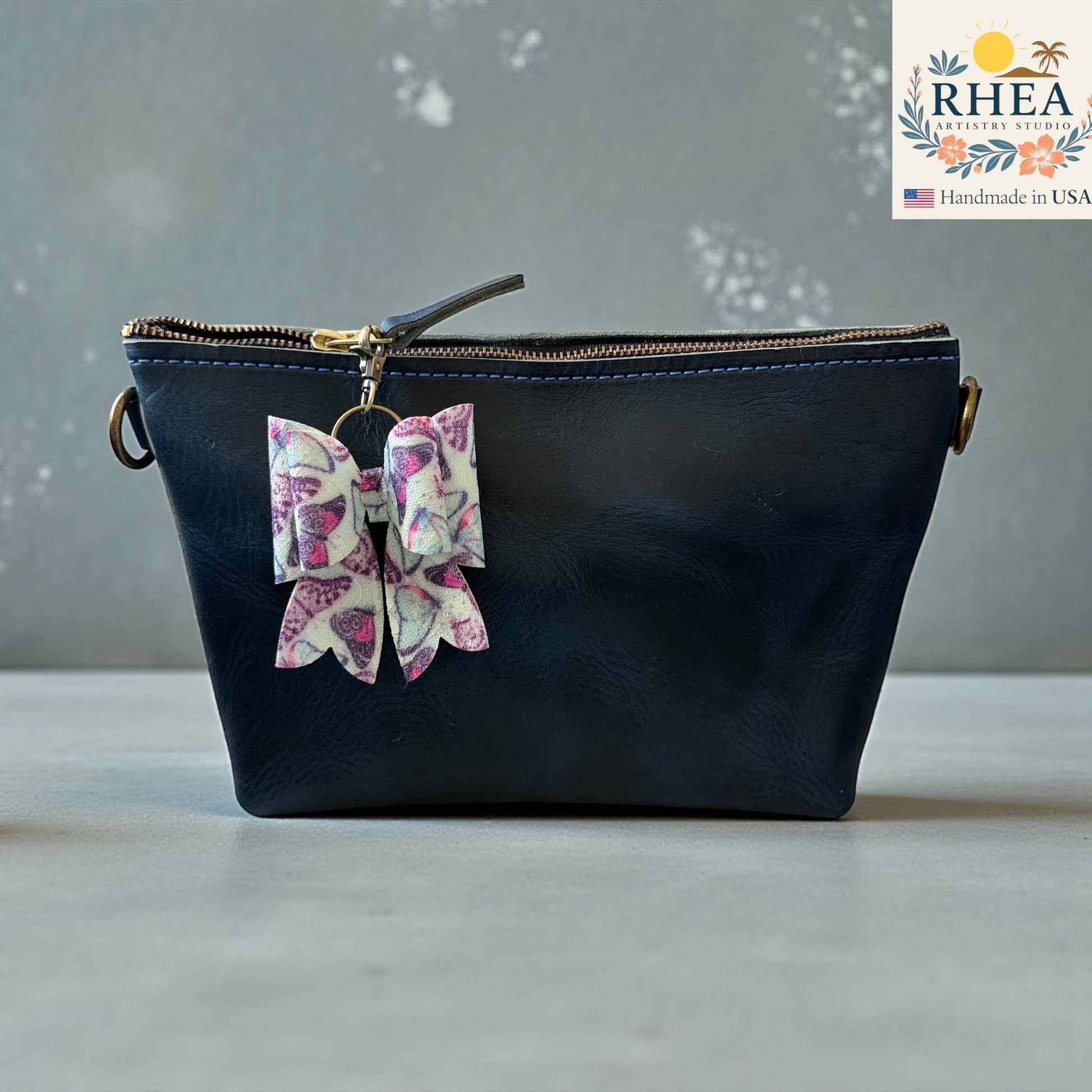 Rhea Artistry Studio handmade double-layer French bow charm with pink and purple butterfly print and brass hardware, clipped to a black leather bag.