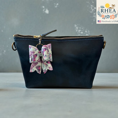 Rhea Artistry Studio handmade double-layer French bow charm with pink and purple butterfly print and brass hardware, clipped to a black leather bag.