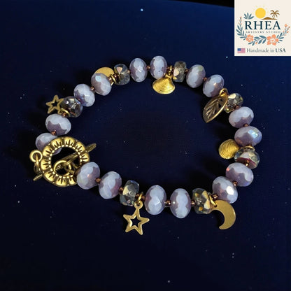 Rhea Artistry Studio handmade bracelet with iridescent glass beads, brass celestial charms (stars, moon, shells, leaf) and floral toggle clasp.