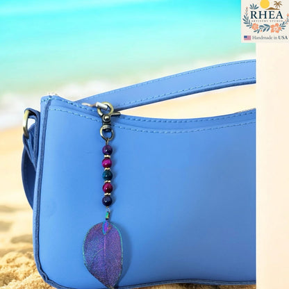 Rhea Artistry Studio celestial cascade bag charm with purple iridescent leaf pendant and gemstone beads on a blue purse.