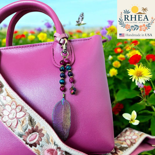 Rhea Artistry Studio Celestial Cascade bag charm with purple, teal, and gold beads and an iridescent leaf pendant.