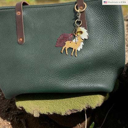 Rhea Artistry Studio Christmas Tales leather charm with a glittery deer, autumn leaves, and bell on a dark green pebbled leather tote bag.