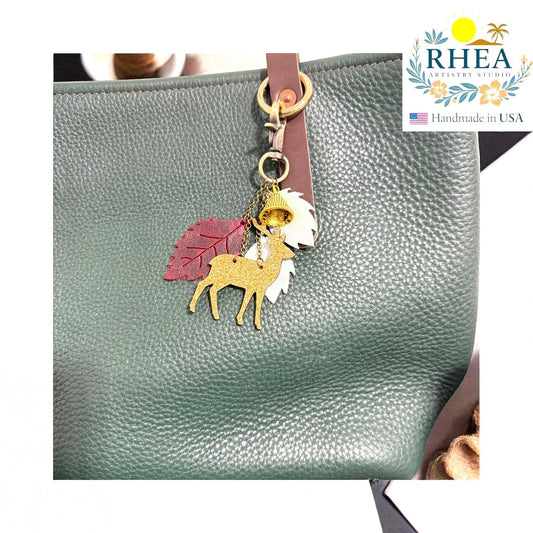 Rhea Artistry Studio handmade leather Christmas charm with gold glitter deer, red leaf, white leaf, and bell on a green pebbled leather bag.
