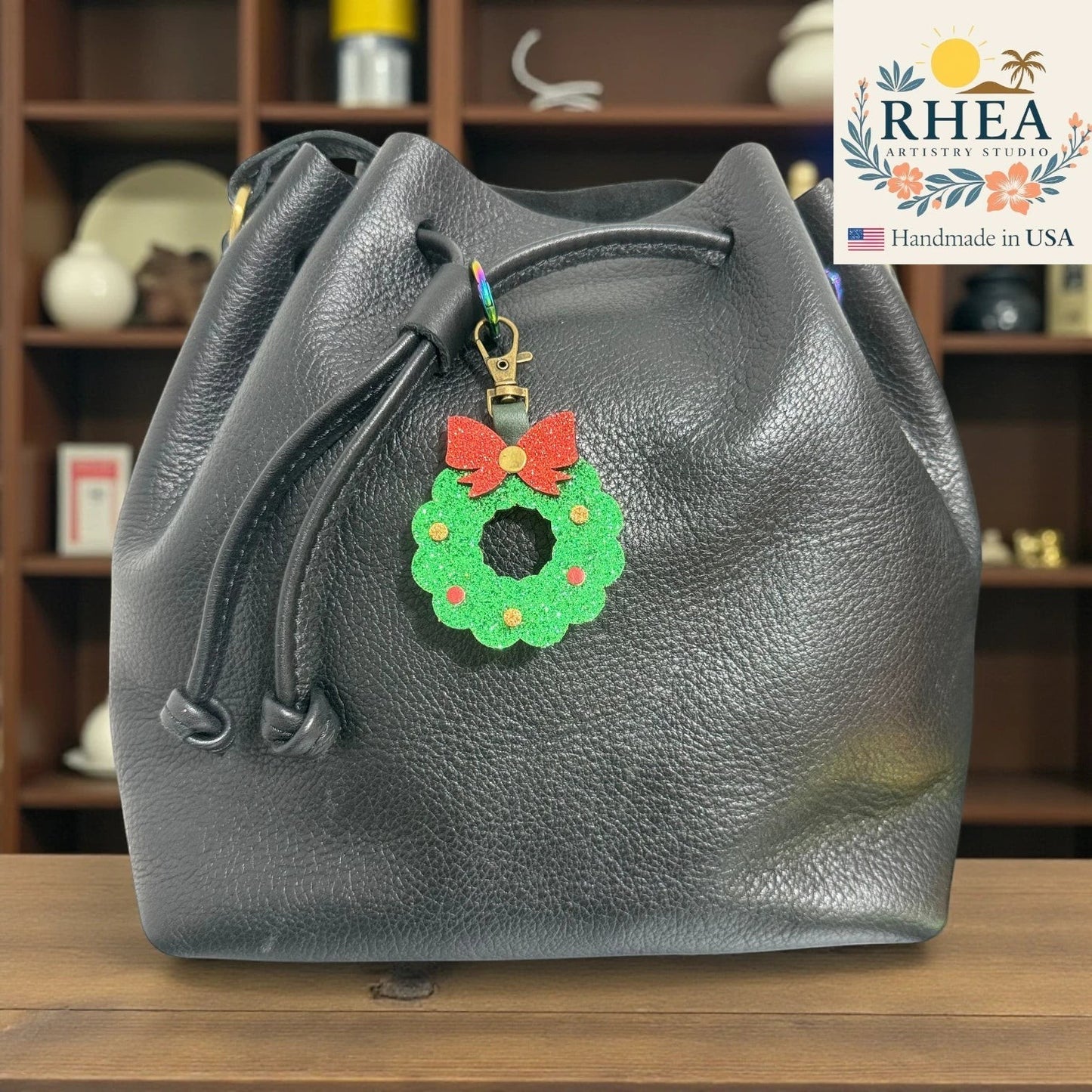 Rhea Artistry Studio handmade glittery green Christmas wreath charm with red bow on black leather bucket bag.