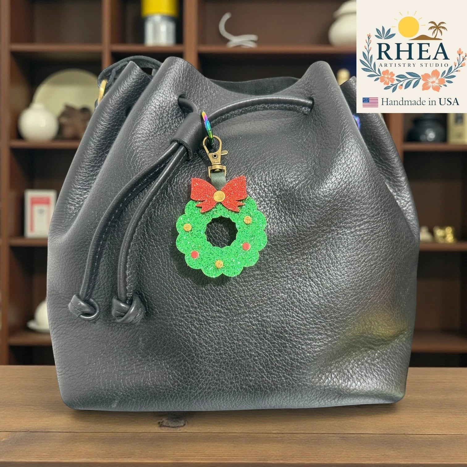 Rhea Artistry Studio handmade glittery green Christmas wreath charm with red bow on black leather bucket bag.