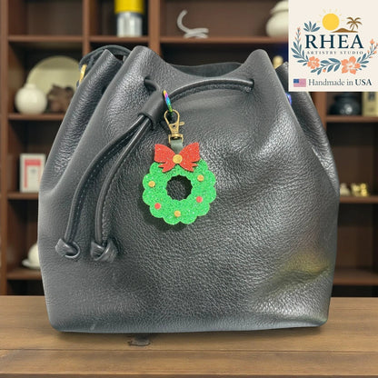 Rhea Artistry Studio handmade glittery green Christmas wreath charm with red bow on black leather bucket bag.