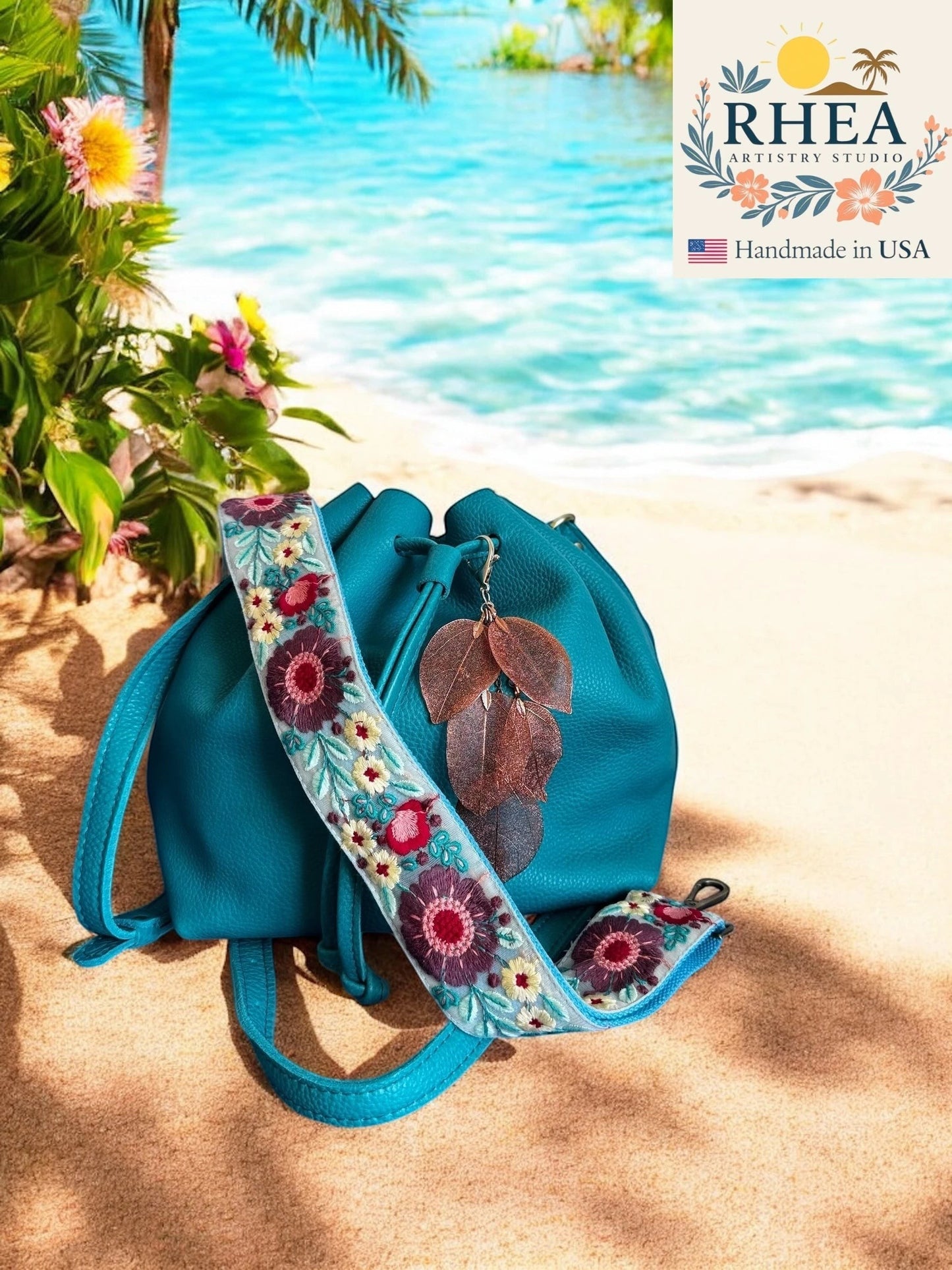 Rhea Artistry Studio handmade copper metallic leaf bag charm with embroidered floral strap on teal bag on beach.