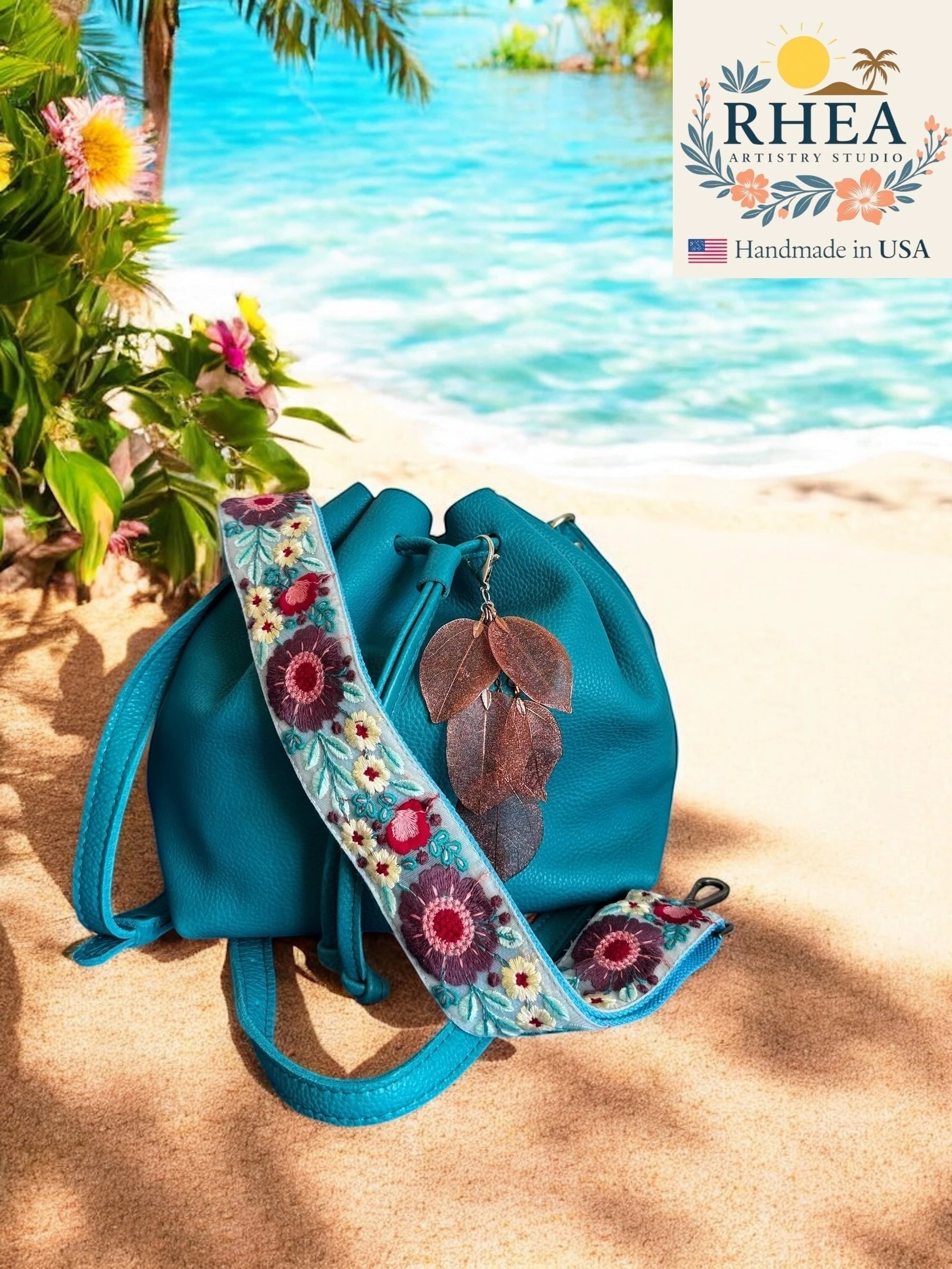 Rhea Artistry Studio handmade copper metallic leaf bag charm with embroidered floral strap on teal bag on beach.