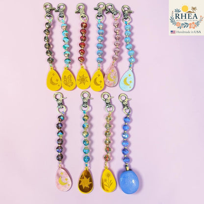 Rhea Artistry Studio artisan bag charms with Czech glass beads, brass accents, and hand-painted pendants featuring celestial and nature motifs.