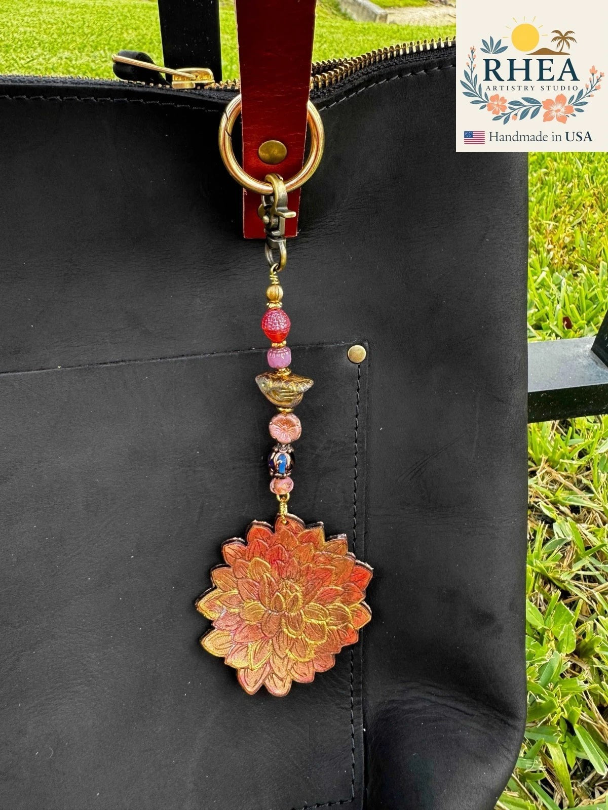 Rhea Artistry Studio handcrafted charm featuring a detailed orange and gold dahlia pendant with colorful beads, attached to a black leather bag.