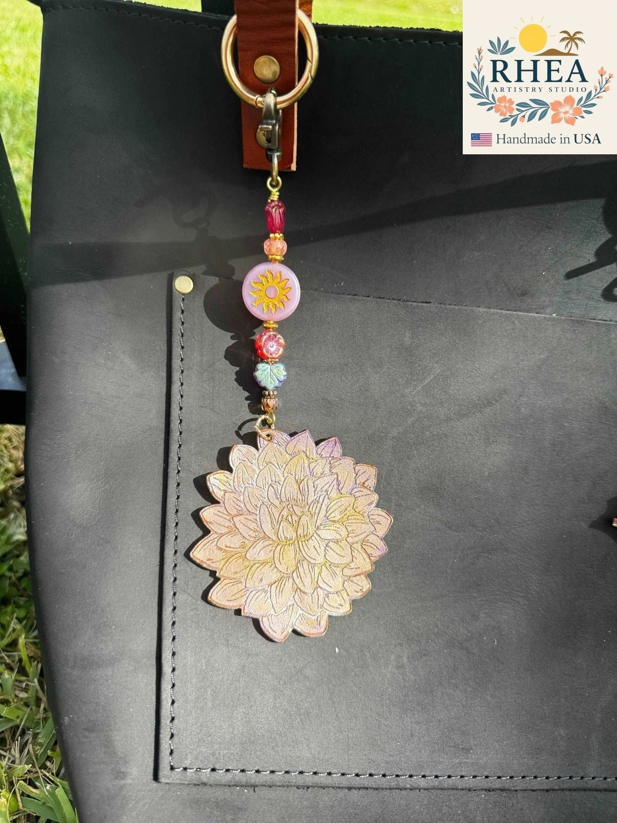 Rhea Artistry Studio Dahlia coin charm with beaded strand featuring a sun charm and colorful glass beads, attached to a black leather bag.