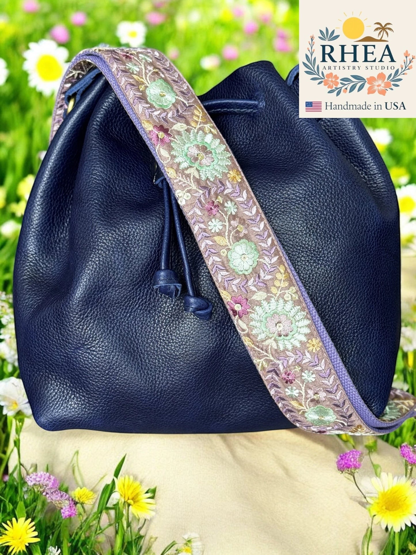 Rhea Artistry Studio navy leather bucket bag with a 2-inch wide Dahlia embroidered floral strap in purple, green, and pink.