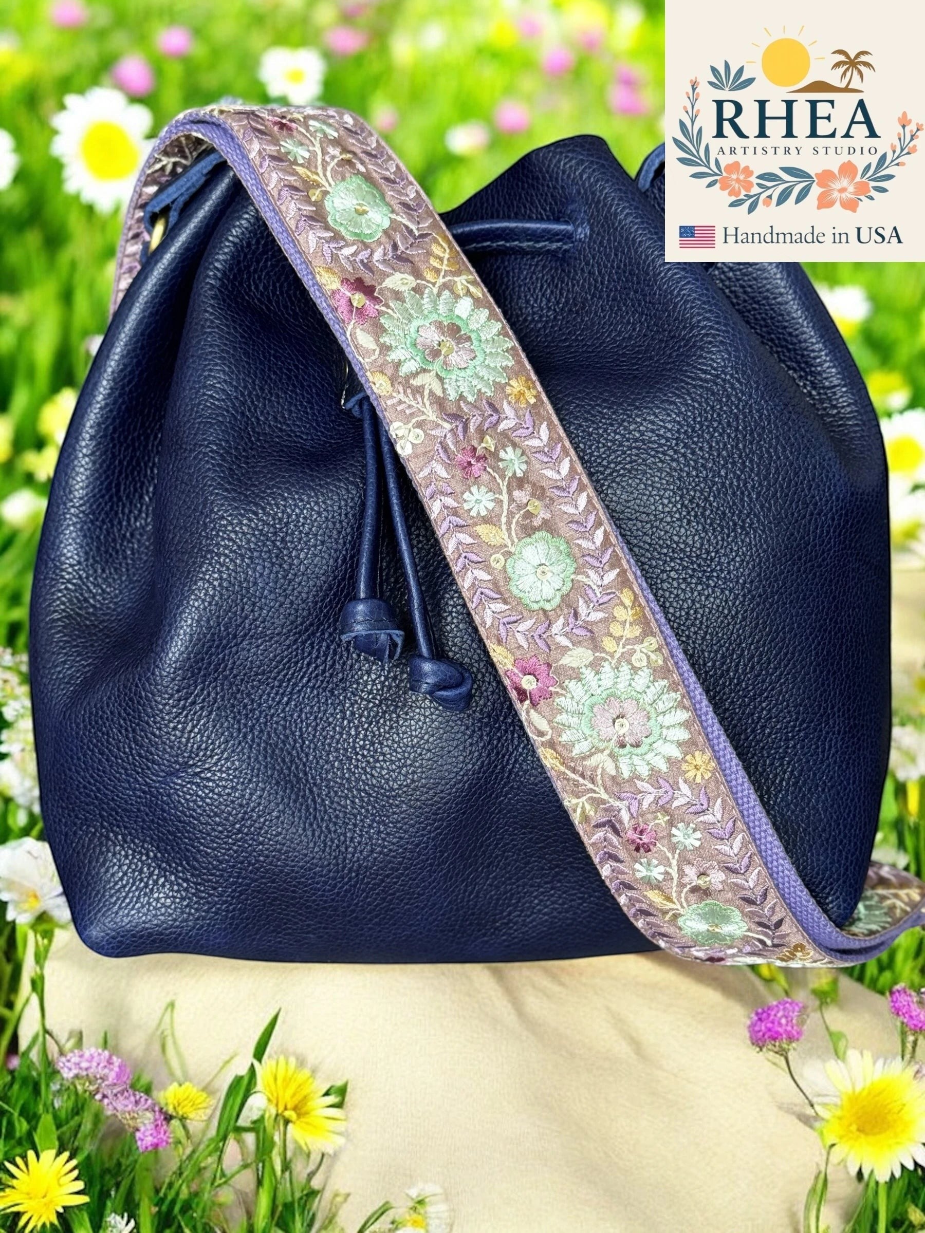 Rhea Artistry Studio navy leather bucket bag with a 2-inch wide Dahlia embroidered floral strap in purple, green, and pink.