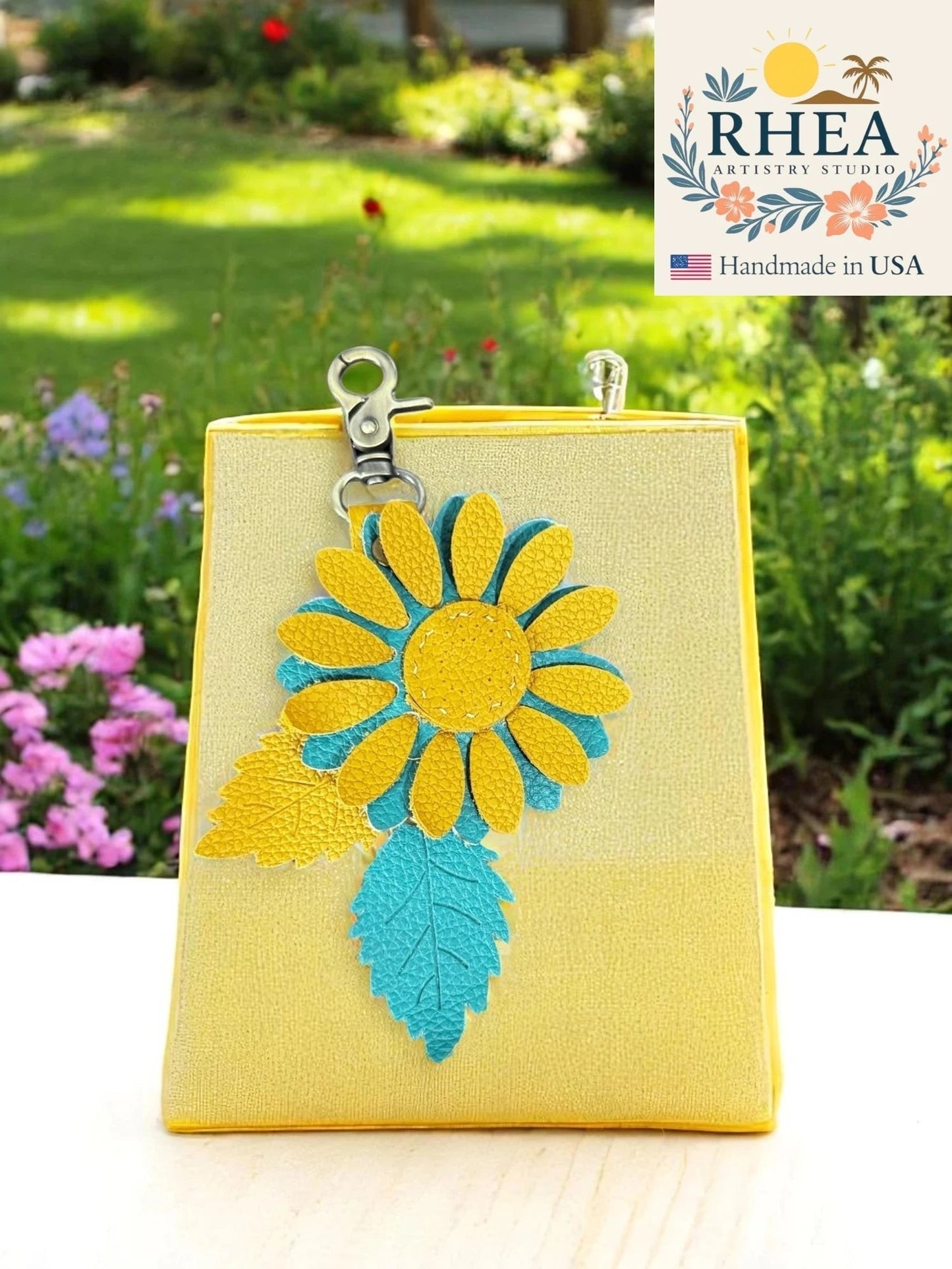 Rhea Artistry Studio yellow leather charm with a blue and yellow daisy and textured leaf detail, attached to a yellow bag with a lobster clasp.
