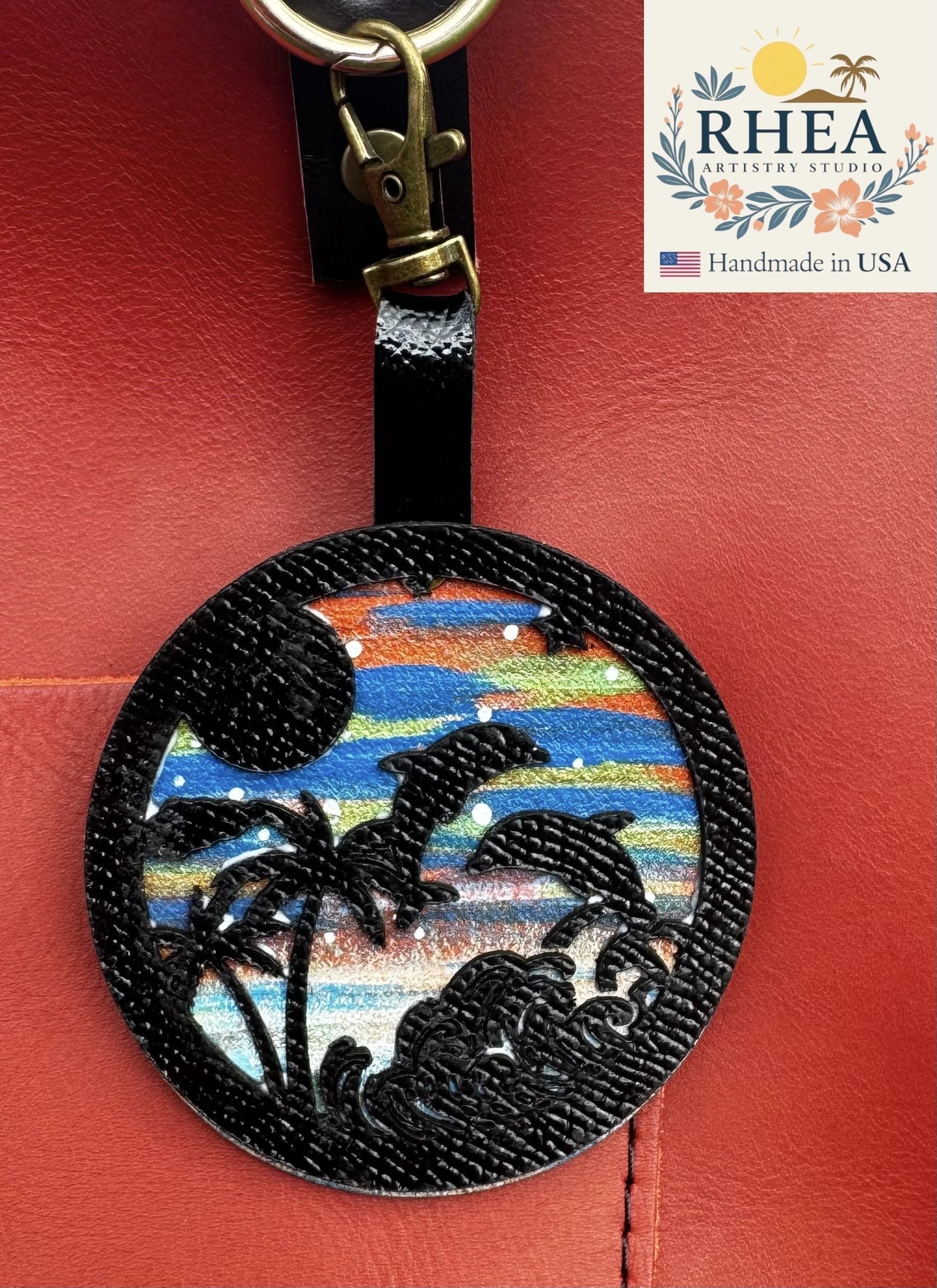 Rhea Artistry Studio hand-painted leather bag charm featuring dolphins, palm trees, and waves on a colorful sunset background. Black leather overlay.