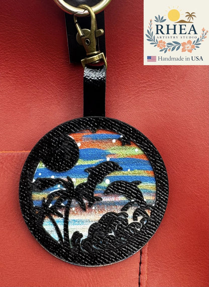 Rhea Artistry Studio hand-painted leather bag charm featuring dolphins, palm trees, and waves on a colorful sunset background. Black leather overlay.