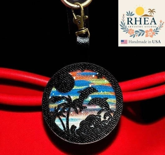 Rhea Artistry Studio hand-painted leather charm featuring dolphins, palm trees, and waves on a sunset sky. Attached with a lobster clasp.