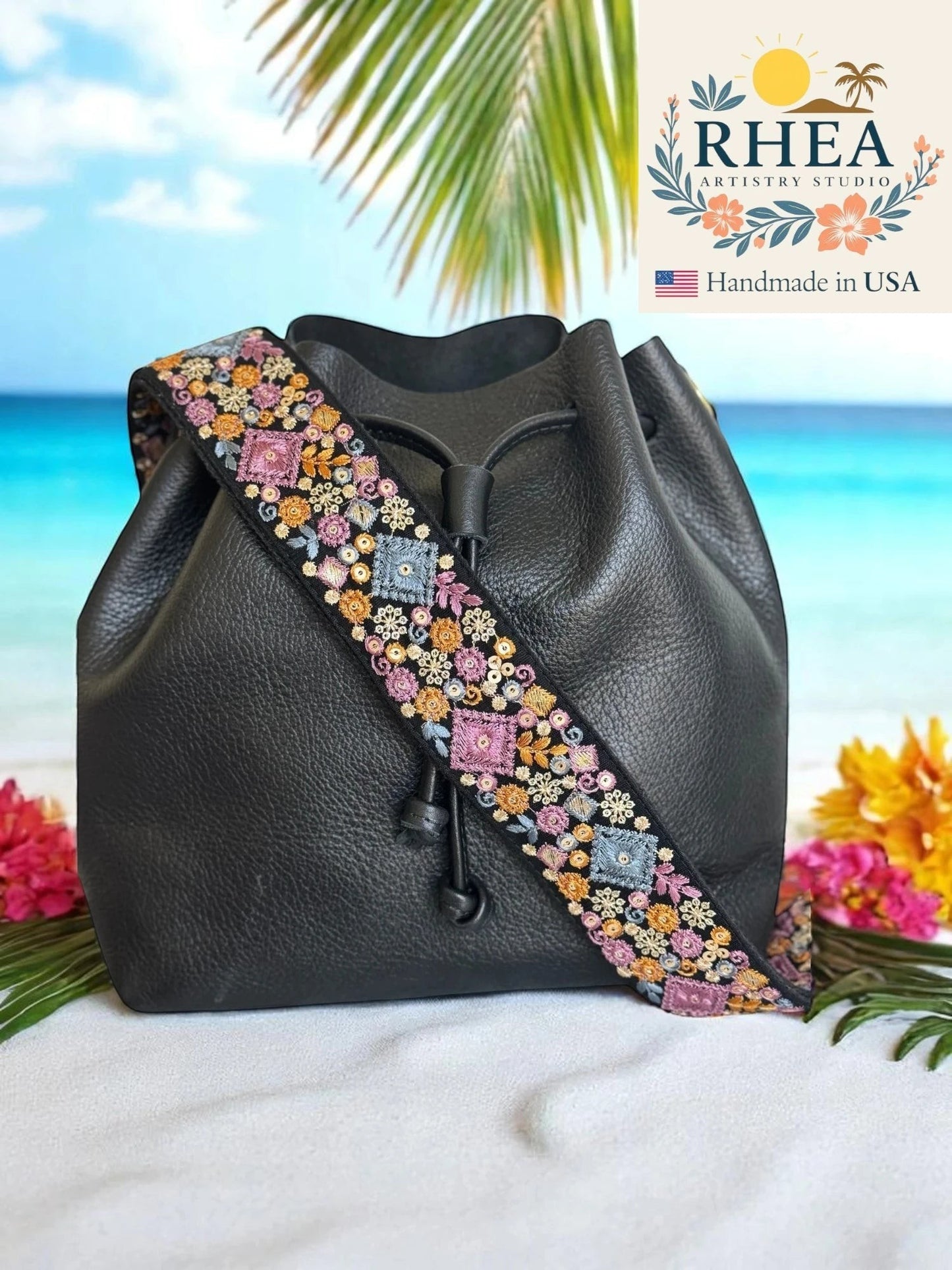 Rhea Artistry Studio black leather bucket bag with a 2-inch wide embroidered strap featuring colorful floral and geometric patterns.