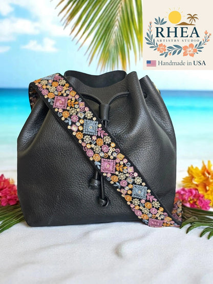 Rhea Artistry Studio black leather bucket bag with a 2-inch wide embroidered strap featuring colorful floral and geometric patterns.