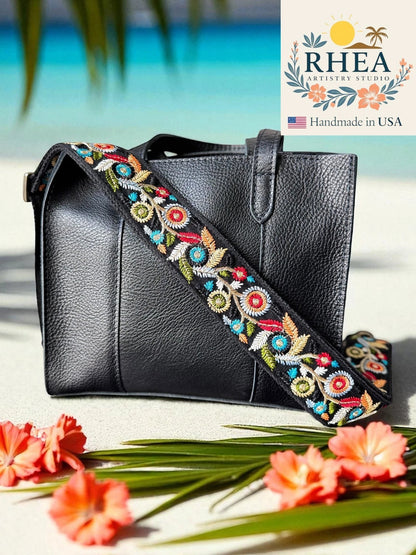 Rhea Artistry Studio black crossbody bag with a 2-inch wide adjustable strap featuring colorful floral embroidery and brushed brass hardware.
