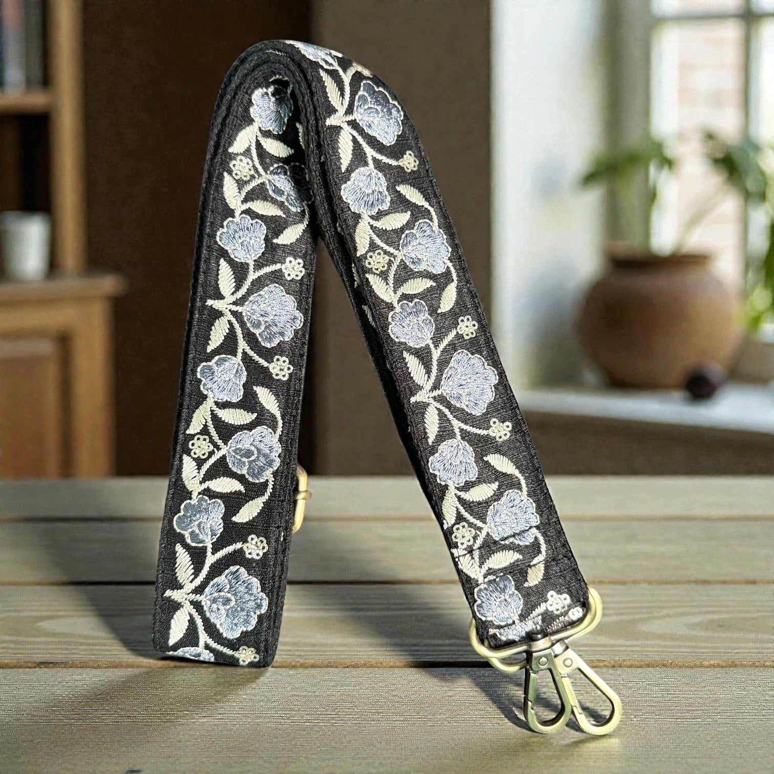Rhea Artistry Studio black crossbody strap with embroidered Sierra Bloom floral pattern in blue and cream, brushed brass hardware.