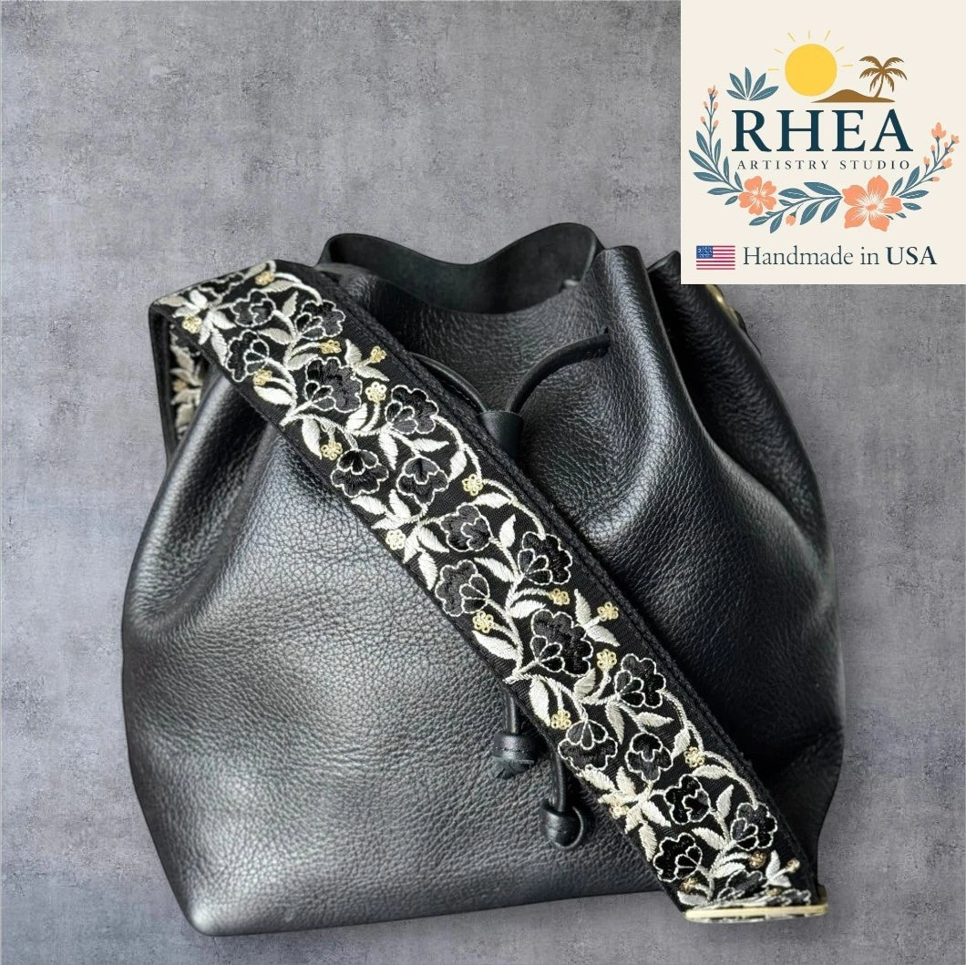 Rhea Artistry Studio 2-inch wide black crossbody strap with white and gold floral embroidery, adjustable length, brushed brass hardware.