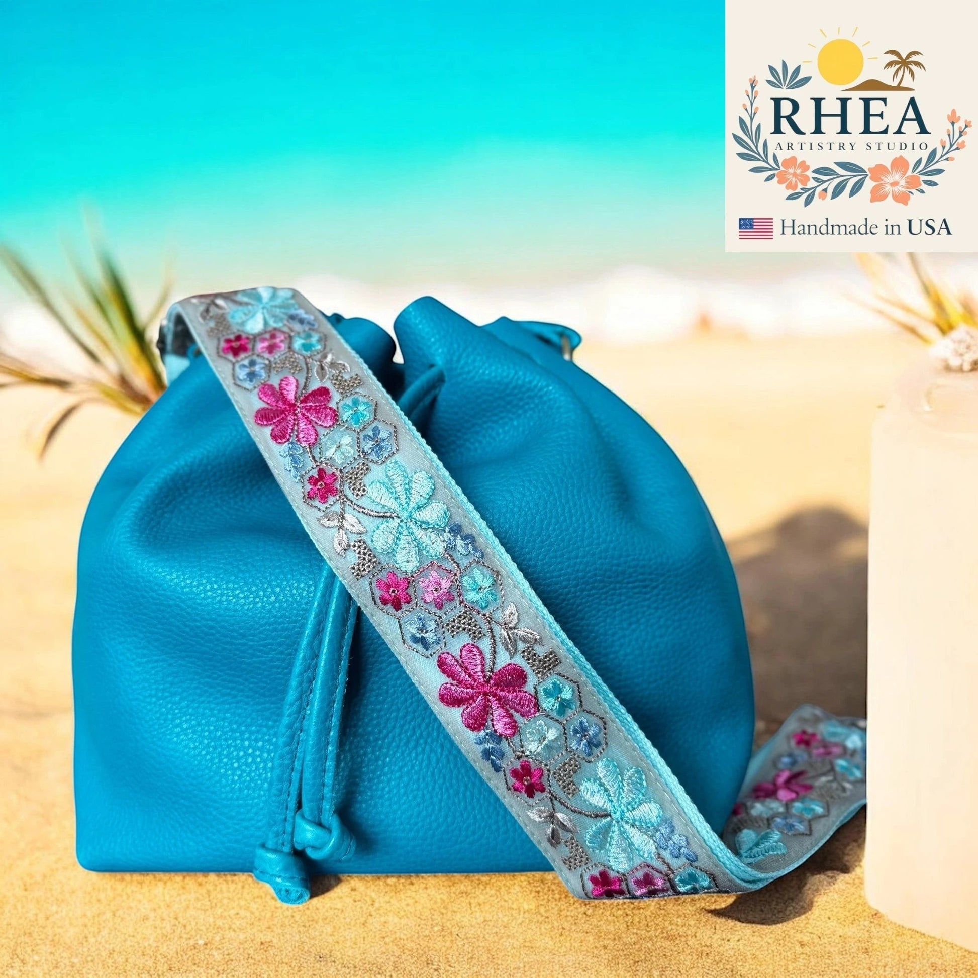 Rhea Artistry Studio 2-inch crossbody strap with floral embroidery in pink, blue, and silver on a light grey background, attached to a blue leather drawstring bag on a sandy beach.