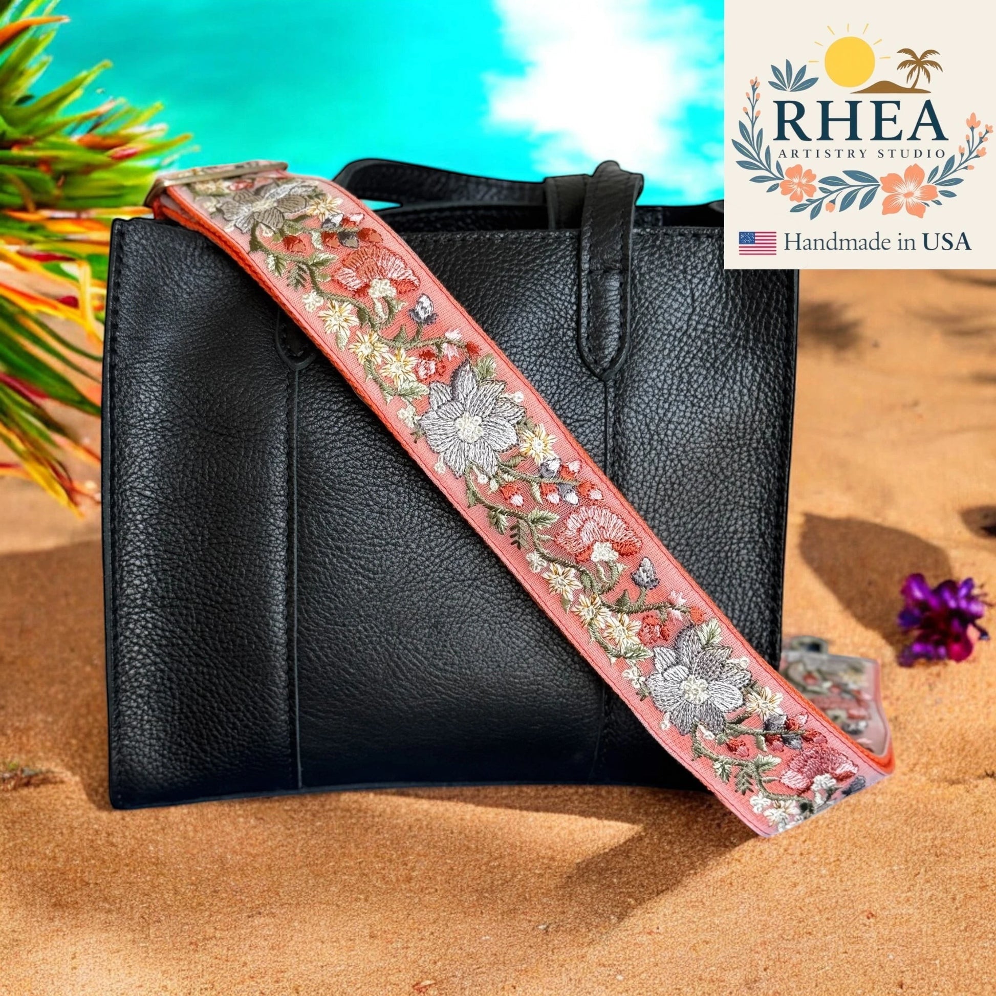 Rhea Artistry Studio coral embroidered fabric crossbody strap with floral design and brushed brass hardware on a black leather tote bag.