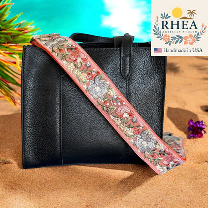 Rhea Artistry Studio coral embroidered fabric crossbody strap with floral design and brushed brass hardware on a black leather tote bag.