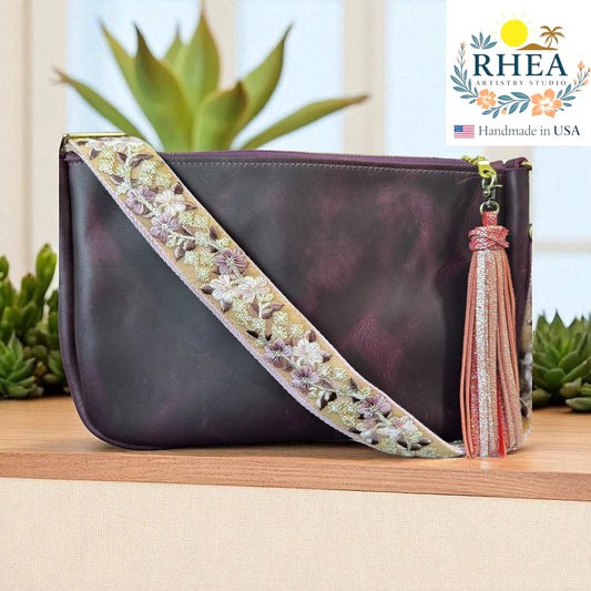 Rhea Artistry Studio crossbody strap with floral embroidery in pink, purple, and green on a tan background, attached to a dark plum leather bag.