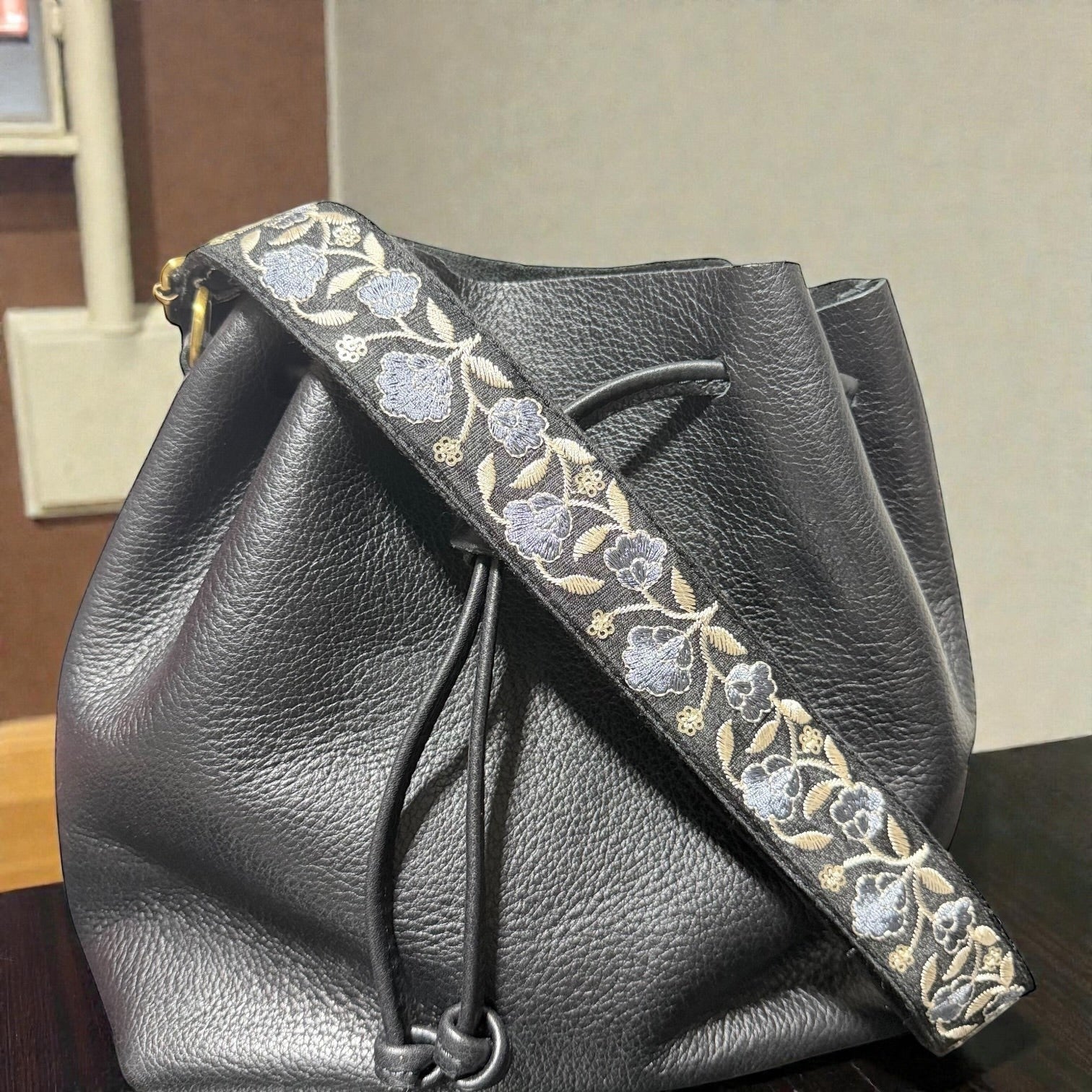 Rhea Artistry Studio Sierra Bloom embroidered crossbody strap with blue and beige floral pattern on black cotton webbing, 1.5-inch wide, adjustable.
