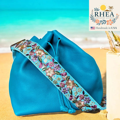 Rhea Artistry Studio 2-inch wide adjustable crossbody strap with floral embroidery in pink, purple, and gold on a light blue background, attached to a blue leather bucket bag on a sandy beach.