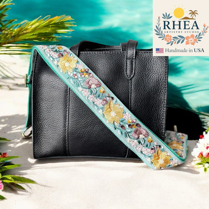 Rhea Artistry Studio light blue embroidered crossbody strap with yellow and pink floral details on a black leather tote bag.