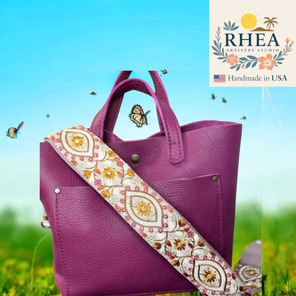 Rhea Artistry Studio 2-inch wide crossbody strap with embroidered floral and geometric patterns in white, gold, and pink on a purple leather handbag.
