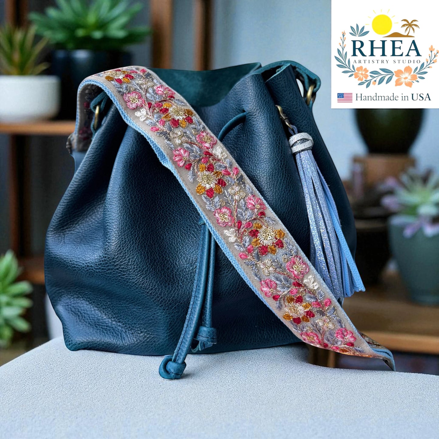 Rhea Artistry Studio embroidered crossbody strap with floral lace ribbon in pink, red, and gold on a grey background, attached to a teal leather bucket bag.