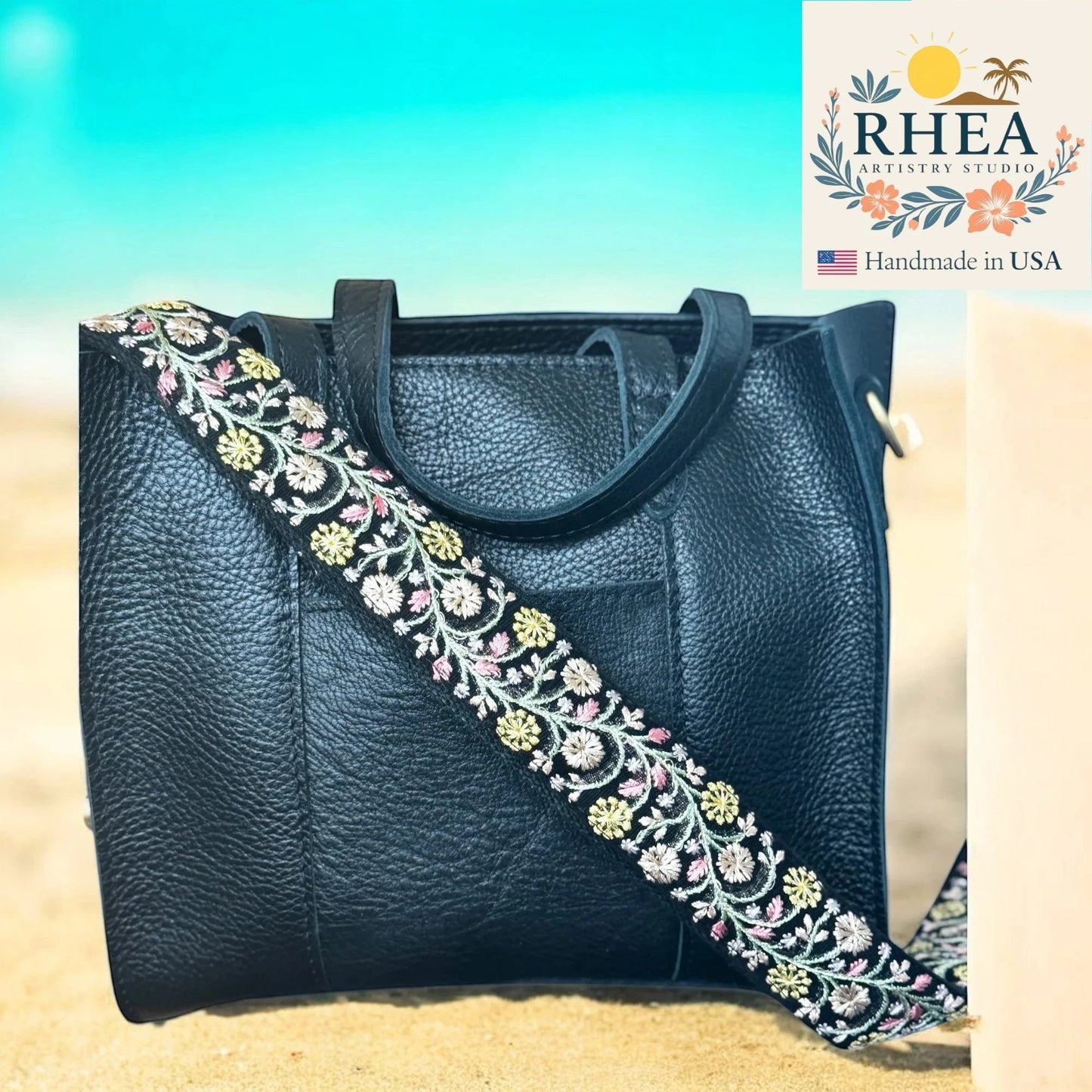 Rhea Artistry Studio embroidered crossbody strap with floral pattern in ivory, pink, and yellow on a black leather bag.