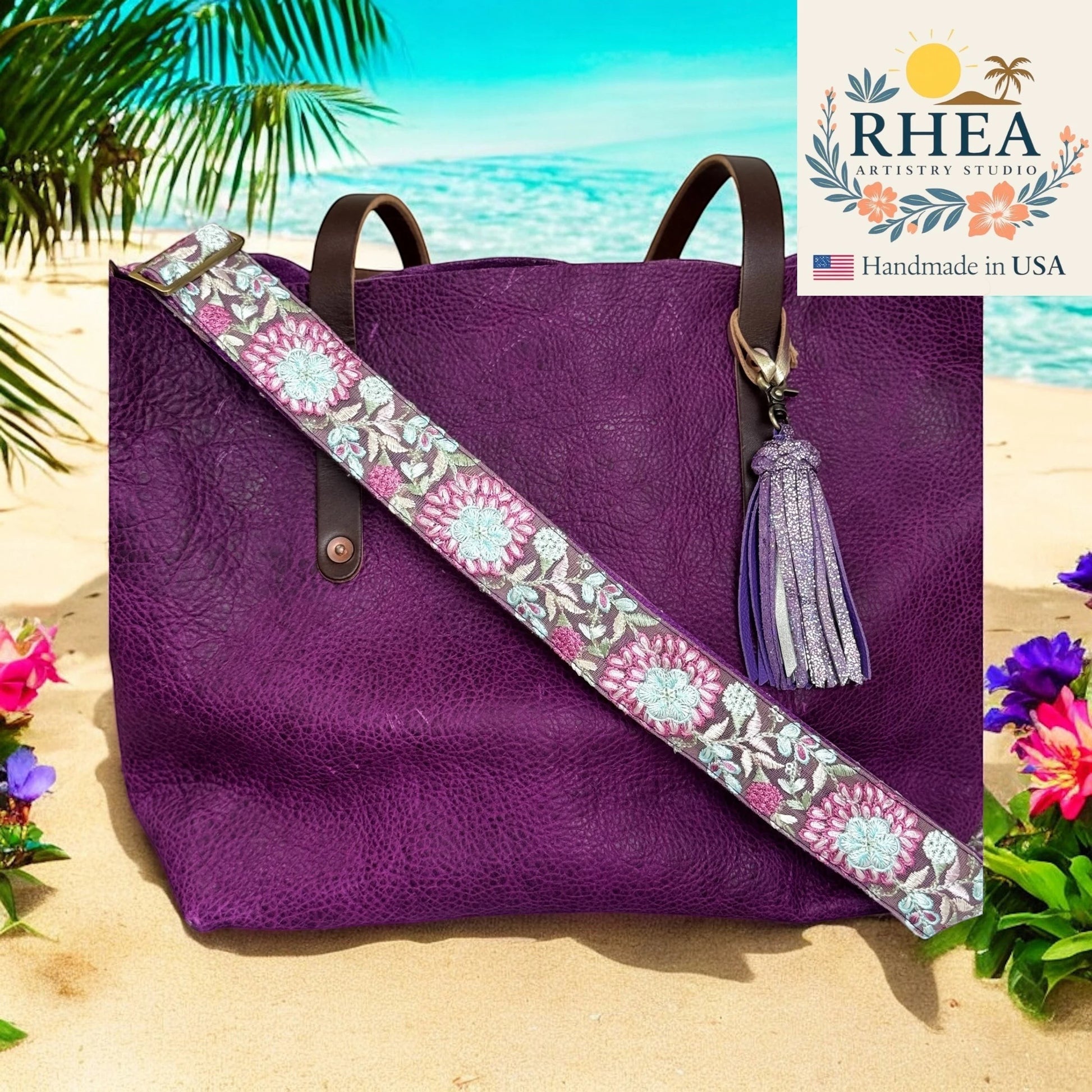 Rhea Artistry Studio purple embroidered crossbody strap with floral detail and brushed brass hardware, shown on a purple leather tote bag on a beach.
